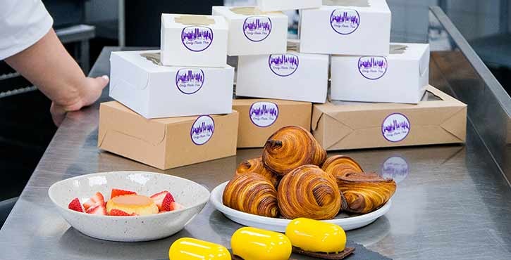 A chef‘s hand reaching for a plate of desserts, including yellow pastries and croissants, with dessert boxes in the background.
