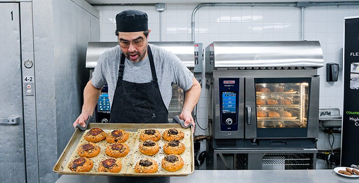 A chef in a black hat and apron holding a tray of freshly baked pastries, with ovens in the background.