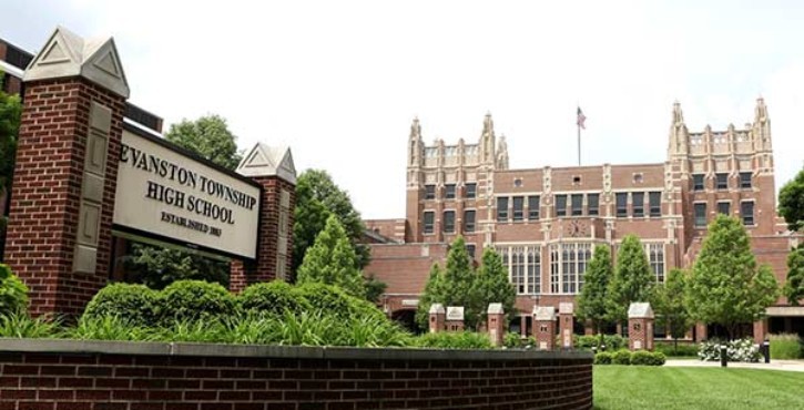 A view of Evanston Township High School's exterior with a sign, lush greenery, and a historic building in the background.