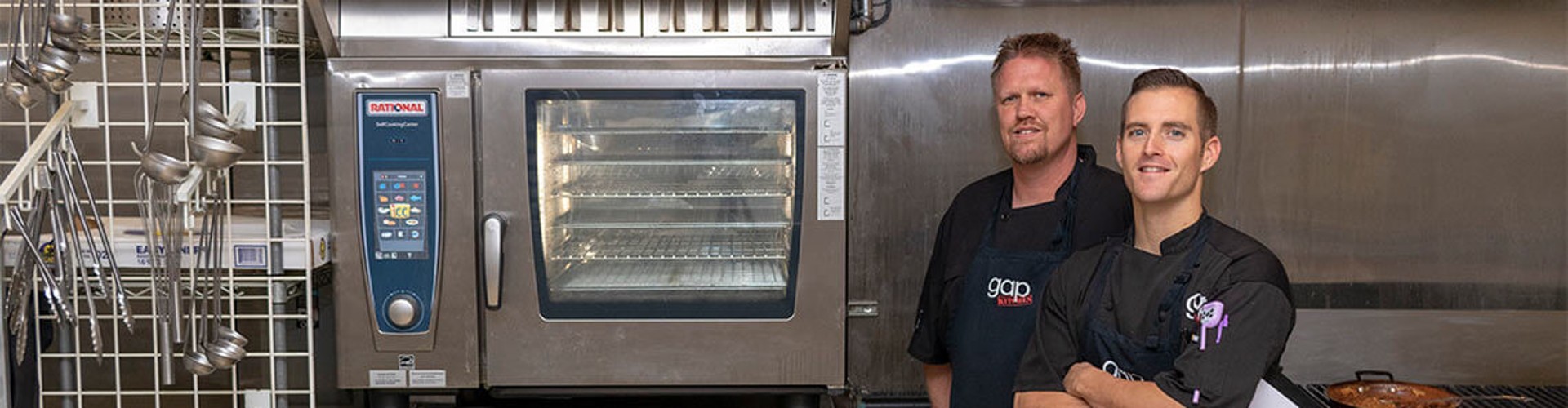Two chefs smiling in a kitchen next to a Rational cooking unit, wearing black aprons with ‘gap‘ logo.
