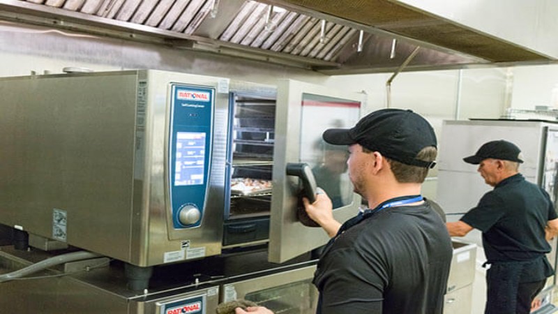 Chef operating a Rational combi oven in a commercial kitchen while another chef observes in the background.