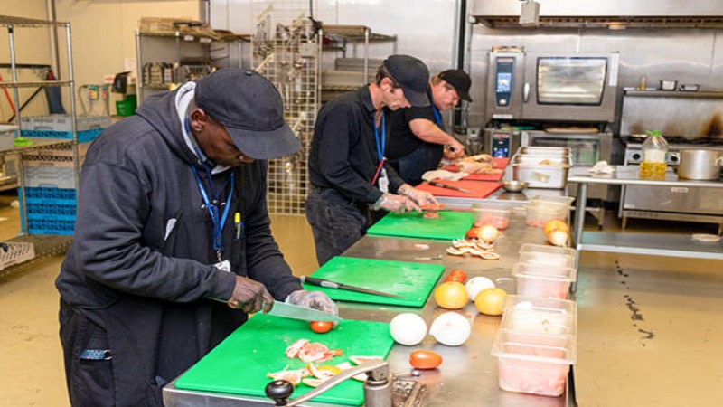 Three chefs preparing vegetables on green cutting boards in a commercial kitchen, wearing black hats and gloves.