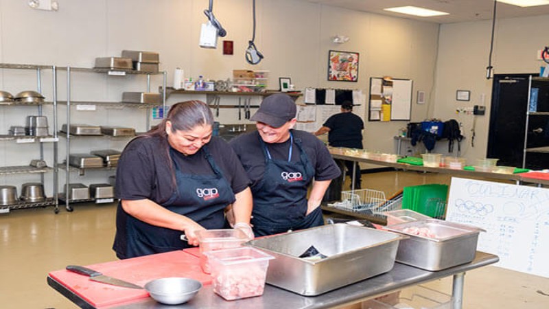 Two chefs preparing food together in a commercial kitchen, with containers and ingredients on the countertop.