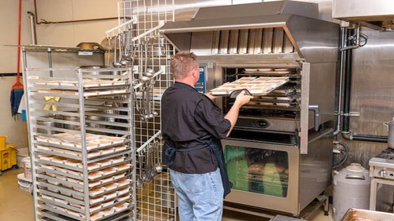 A chef loading trays of prepared meals into a combi oven in a commercial kitchen, with racks of food in the background.
