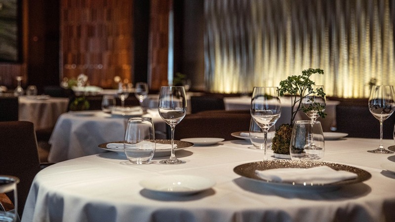 Elegant dining setup at a restaurant with white tablecloths, wine glasses, and plates, featuring a subtle ambiance highlighted by soft lighting.