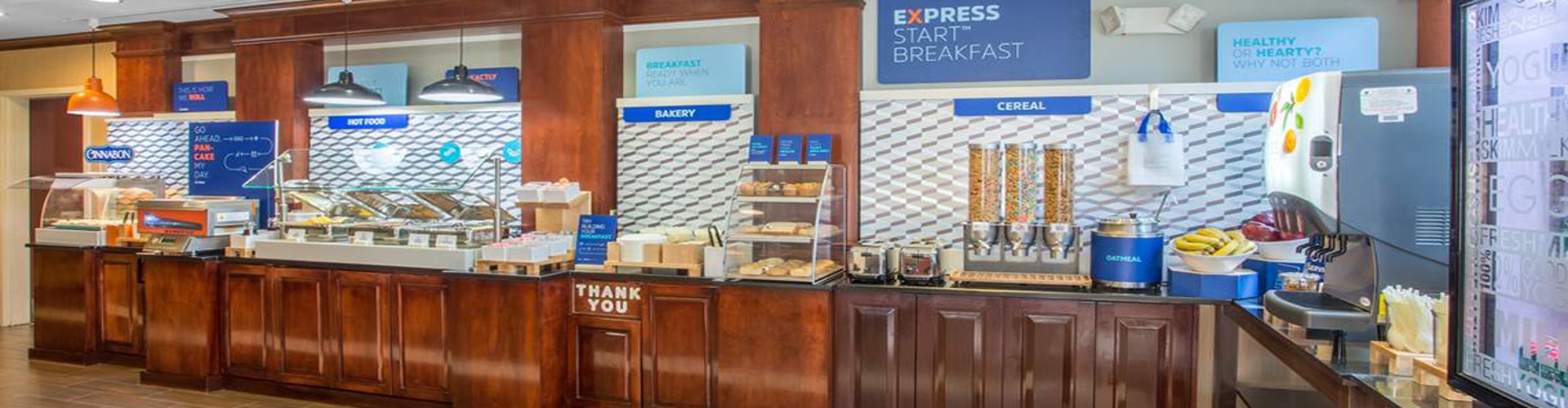 Interior of a Holiday Inn Express breakfast area with buffet counters offering various foods under signs for ‘Express Start Breakfast‘ and ‘Healthy Options‘, and a ‘Thank You‘ placard displayed.