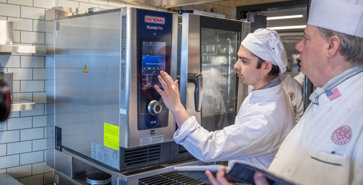 Chef operates a RATIONAL iCombi Pro combi oven touchscreen while another chef observes.