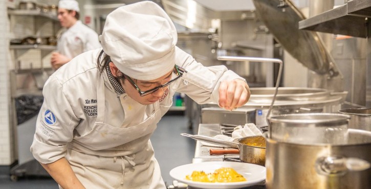 Culinary student carefully seasons a plated dish in a professional kitchen.