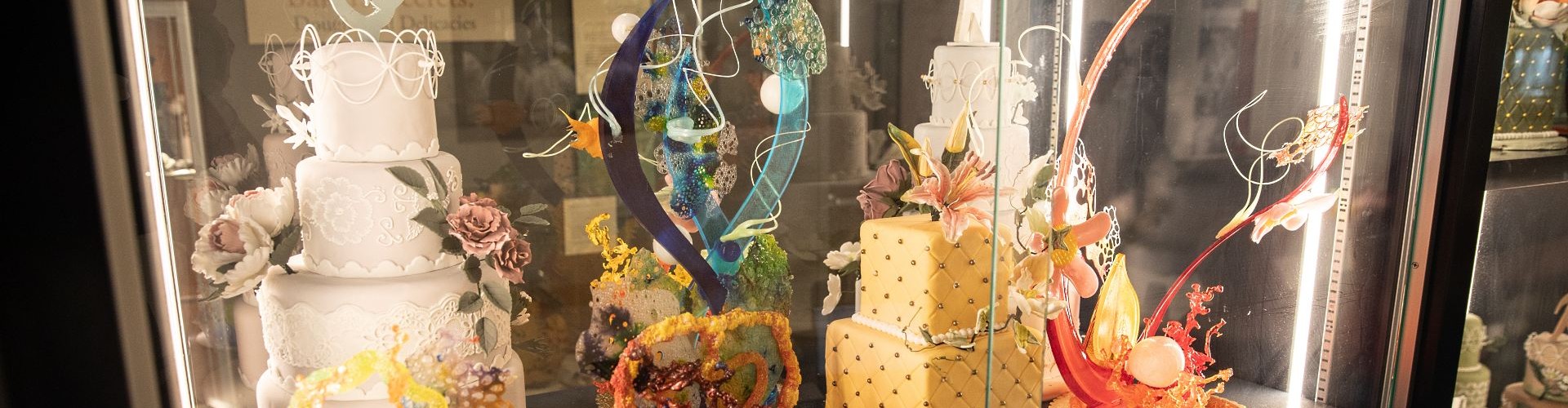 Elaborately decorated tiered cakes and colorful sugar sculptures displayed in a glass case.