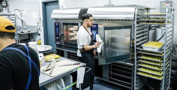 Chef opening a RATIONAL iCombi Pro combi oven beside a rack of prepared trays in a commercial kitchen.