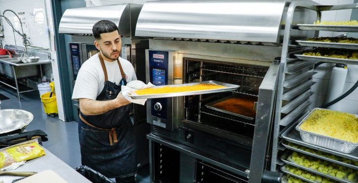 Chef removing a tray from a RATIONAL iCombi Pro combi oven beside a rack of prepared food trays.