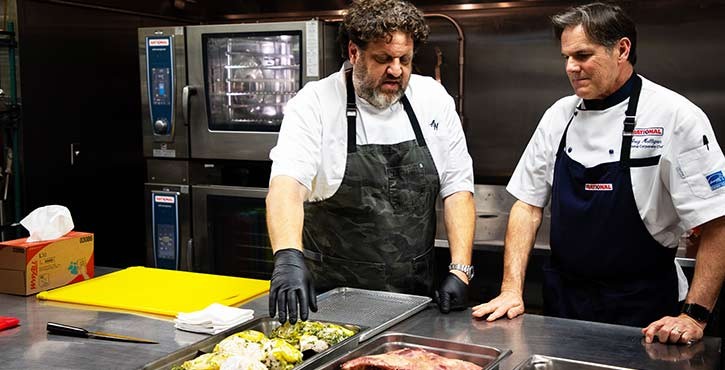 Two chefs discuss the preparation of dishes in a professional kitchen, with ingredients on the counter.