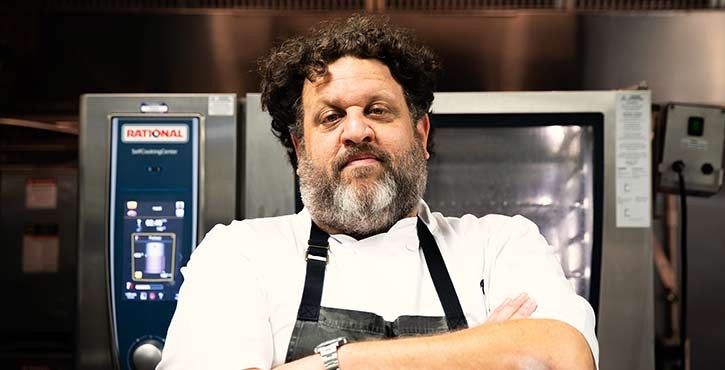 A confident chef stands with arms crossed in a kitchen, looking directly at the camera.