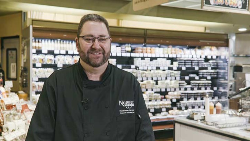A person in a black chef‘s uniform, labeled ‘Nugget Market,‘ smiling in front of a well-stocked cheese counter in a grocery store.