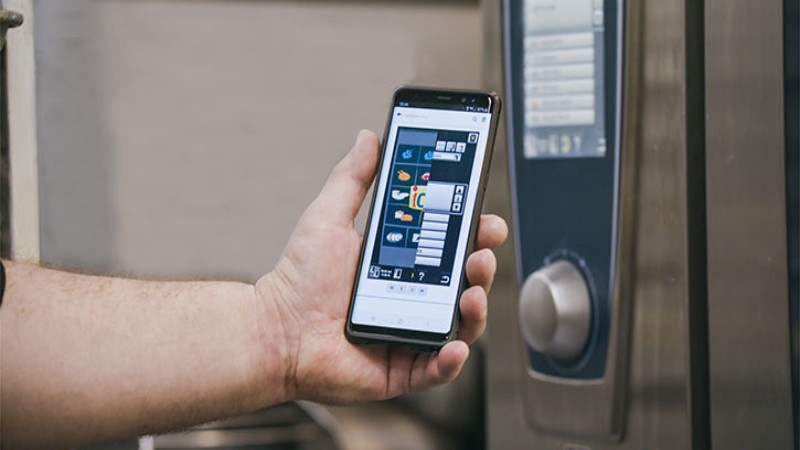 One person uses a smartphone to control the RATIONAL appliance. Colorful icons and digital buttons are displayed on the screen.