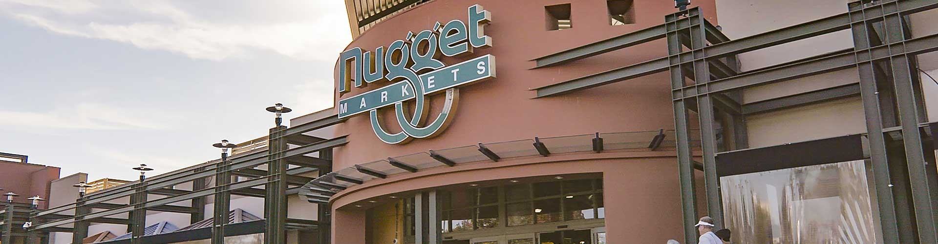 Exterior view of Nugget Markets with a prominent sign, featuring pedestrians and a parked taxi near the entrance under a clear sky.