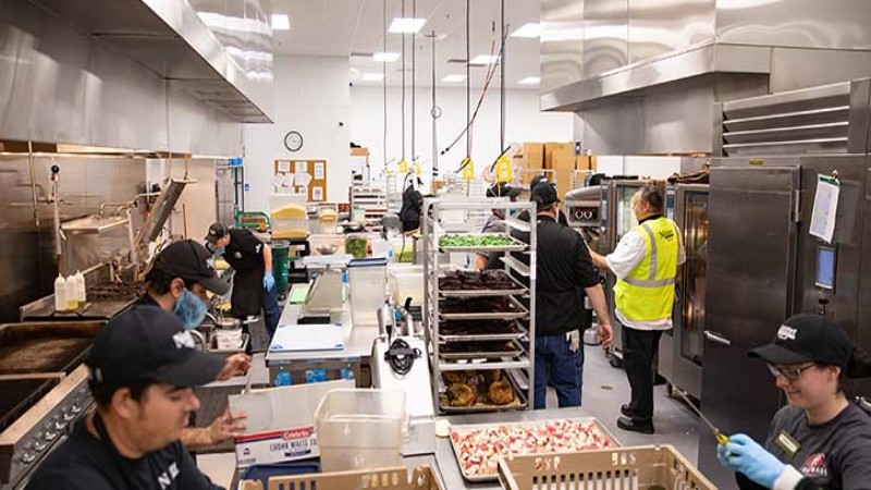 Several workers are busy in a large, industrial kitchen setting, handling food preparation and cleaning tasks at different stations.