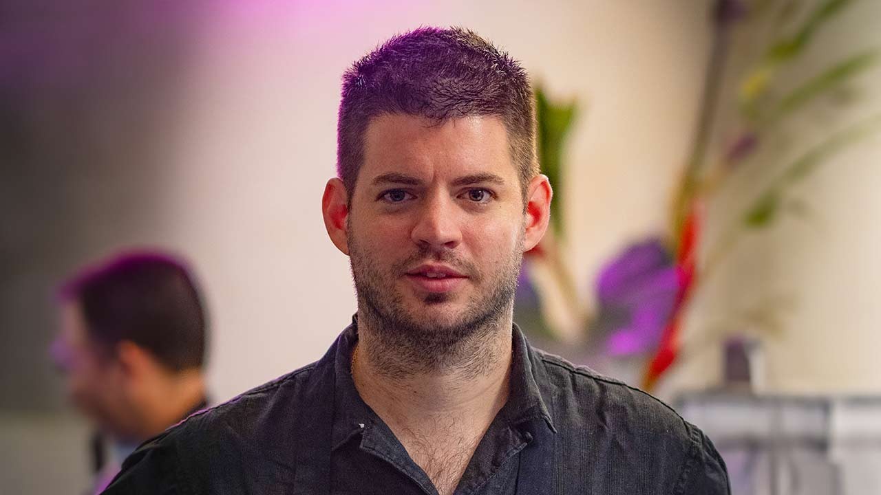 A close-up portrait of a young man with short hair and a stubbled beard, smiling at the camera in a colorful setting.