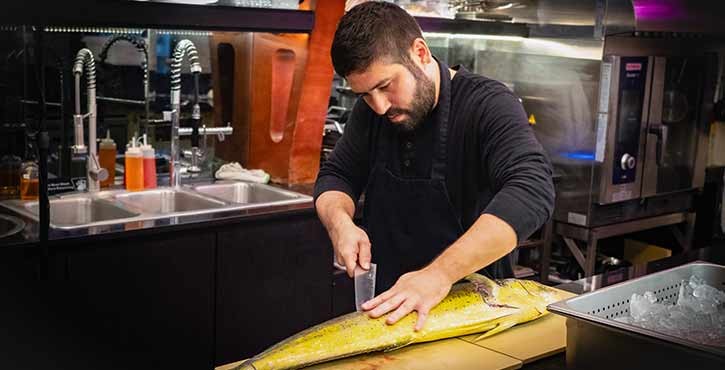 A chef skillfully fillets a large fish on a cutting board in a professional kitchen, with kitchen tools visible in the background.