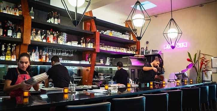 A lively restaurant kitchen with staff working behind the bar and cooking area, showcasing a vibrant atmosphere and a well-stocked shelf.