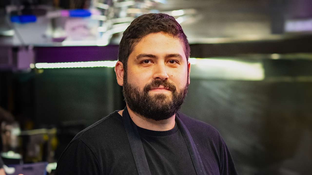 A close-up portrait of a chef with a full beard, wearing a black shirt and apron, smiling confidently in a professional kitchen.