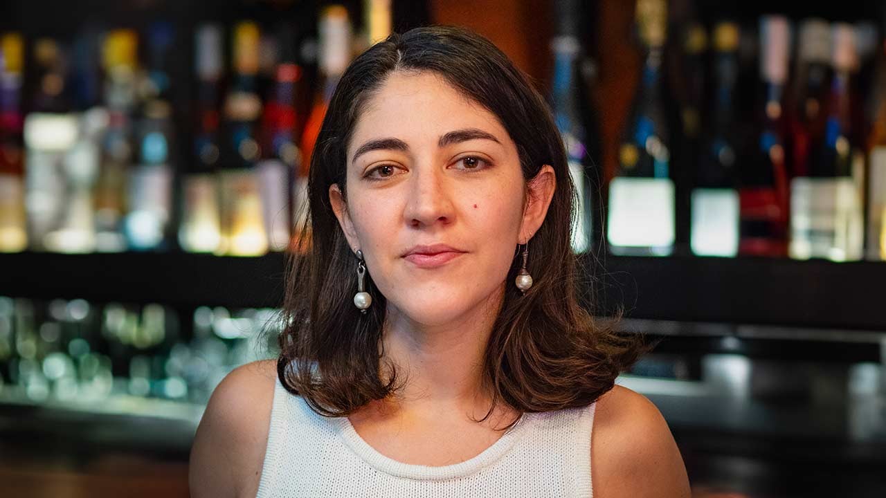 A close-up portrait of a woman with shoulder-length hair, wearing a sleeveless top and pearl earrings, smiling softly against a backdrop of bottles.