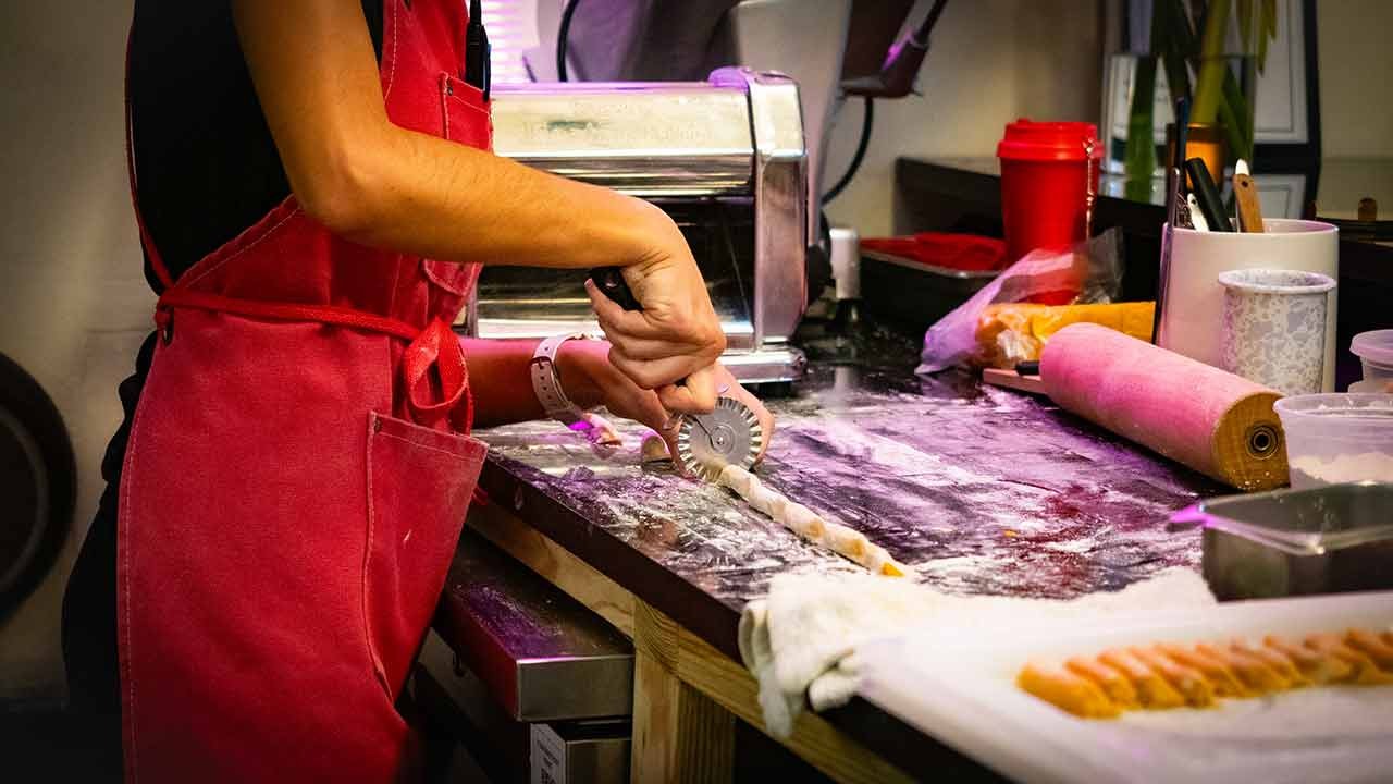 A chef in a red apron uses a pastry wheel to cut rolled-out dough on a floured countertop, surrounded by kitchen tools.