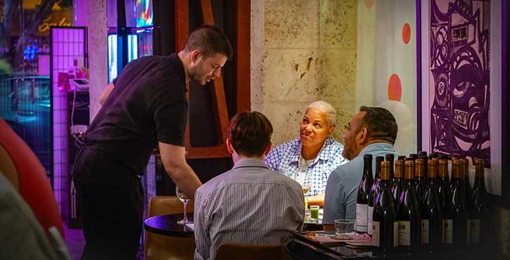 A waiter serves drinks to a table of guests in a vibrant restaurant setting, with colorful lighting and wine bottles in the foreground.