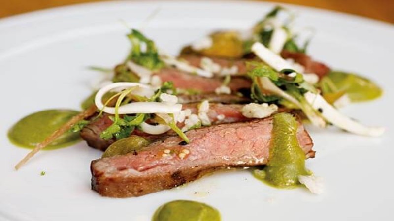 Sliced steak topped with herbs and garnishes, served with green sauce on a white plate.