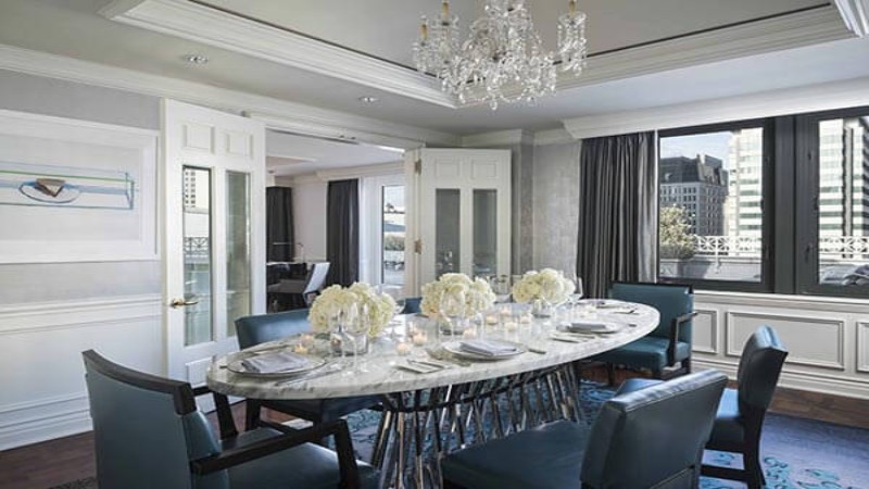 Elegant dining room featuring a glass-topped oval table with blue chairs, set for six. Chandelier overhead, large windows with city view, and tasteful art. Stylish and contemporary decor.