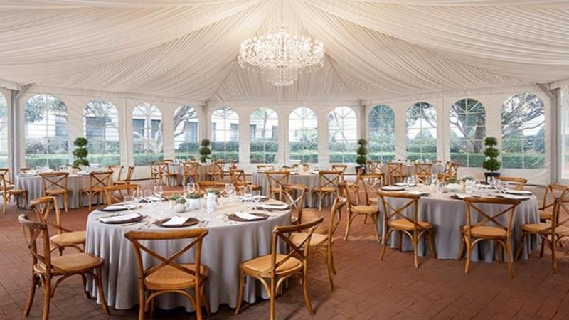 Elegant event space under a large tent with draped white fabric ceiling and chandeliers, featuring round tables set with white tablecloths, fine china, and surrounded by wooden chairs. Large windows show a garden view.