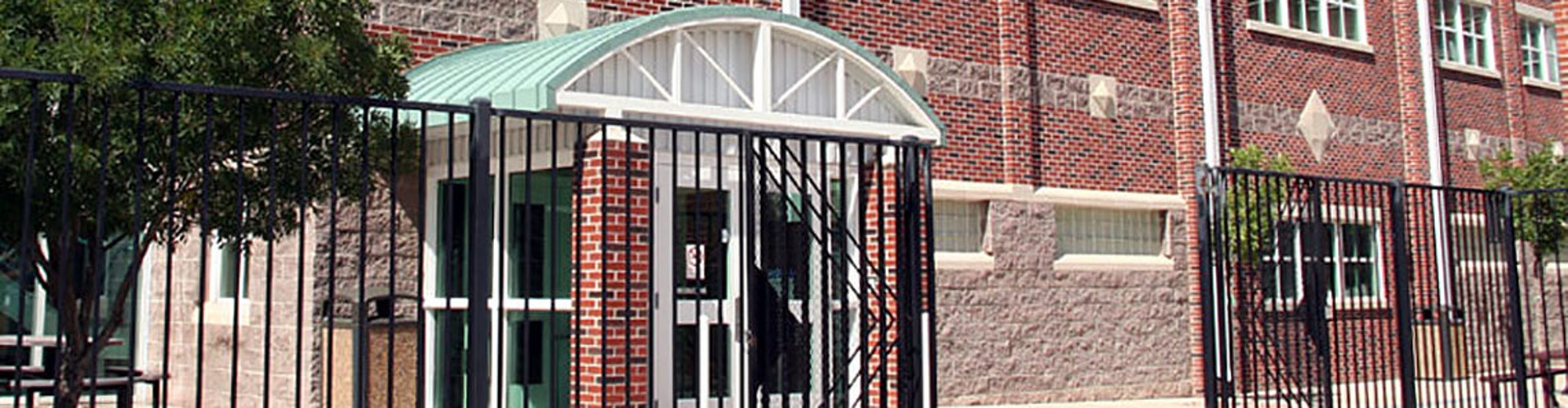 Exterior view of a traditional red brick school building with a gated entrance and arched doorway under a clear sky.