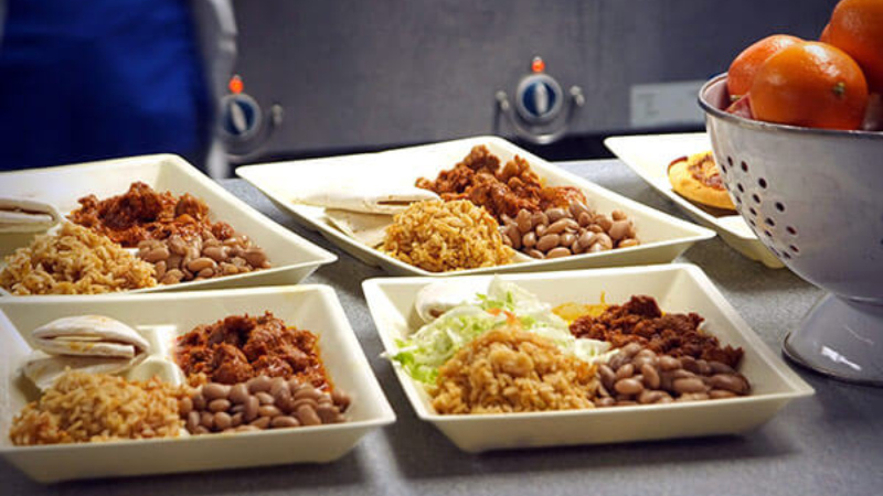 Several trays of food containing rice, beans, and chili, displayed on a table with oranges in a colander in the background.
