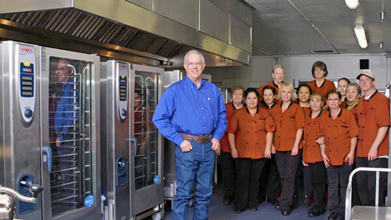 Group of workers in uniform standing in a commercial kitchen with large RATIONAL ovens, led by an individual in a blue shirt smiling at the camera.