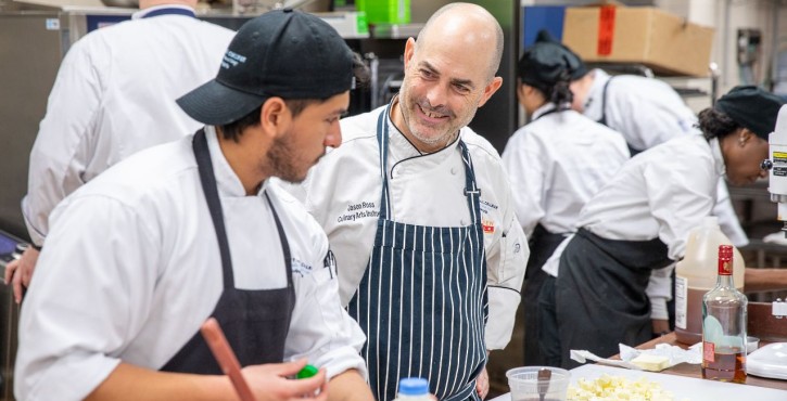 Culinary instructor speaking with a chef as students prepare food in a commercial kitchen.