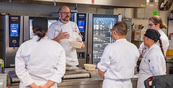 Chef instructing culinary students beside RATIONAL iCombi Pro combi ovens in a commercial kitchen.