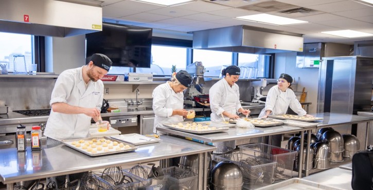 Four culinary students prepare trays of pastries at stainless steel worktables in a commercial kitchen.