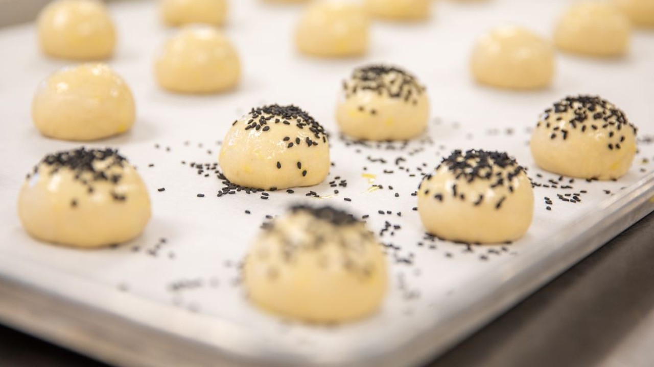 Unbaked rolls topped with black sesame seeds arranged on a baking sheet.