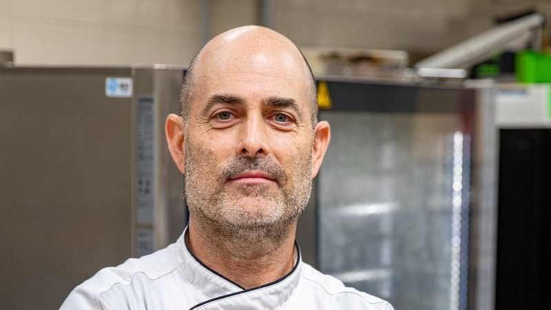 Culinary instructor in a white chef’s coat stands in a professional teaching kitchen.