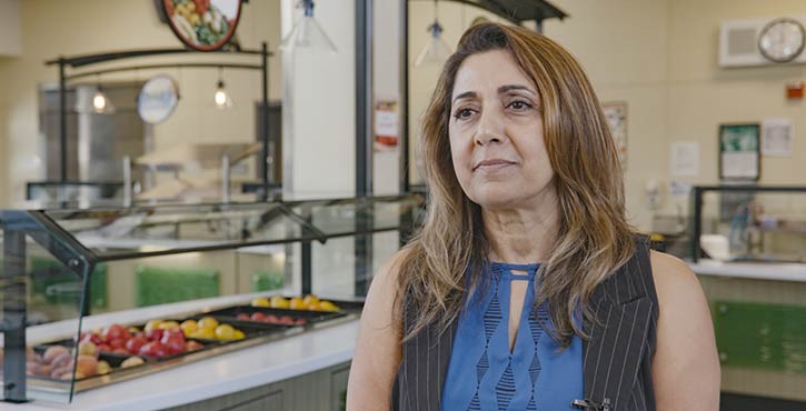 A portrait of Sheri Nielsen standing in a cafeteria setting, wearing a blue blouse, with a salad bar in the background.