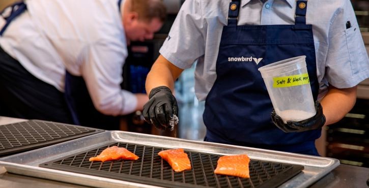 Chef seasoning salmon fillets on trays in a commercial kitchen while another chef works in the background.