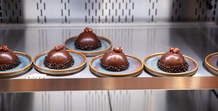 Chocolate dome desserts plated on small dishes on a stainless-steel kitchen shelf.