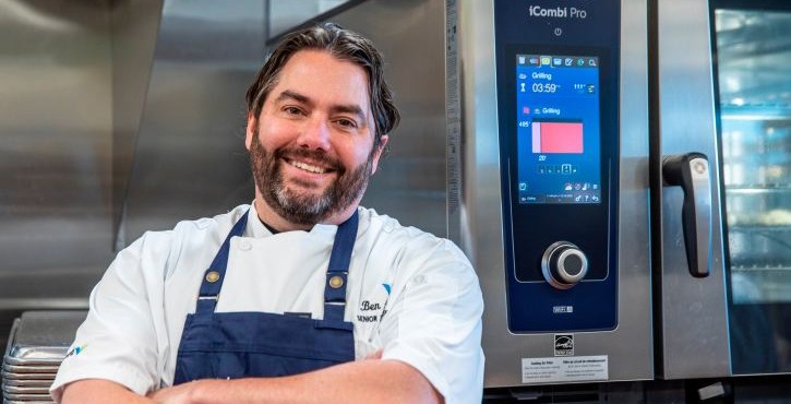 Smiling chef standing beside a RATIONAL iCombi Pro combi oven in a commercial kitchen.