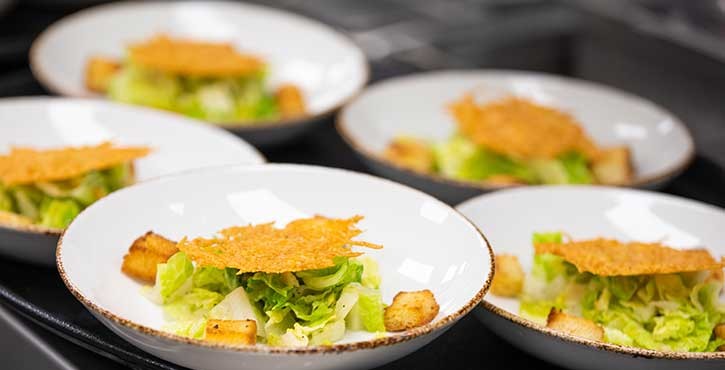 Plates of Caesar salad with crisp croutons and Parmesan crisps, arranged in a row on a dark countertop.
