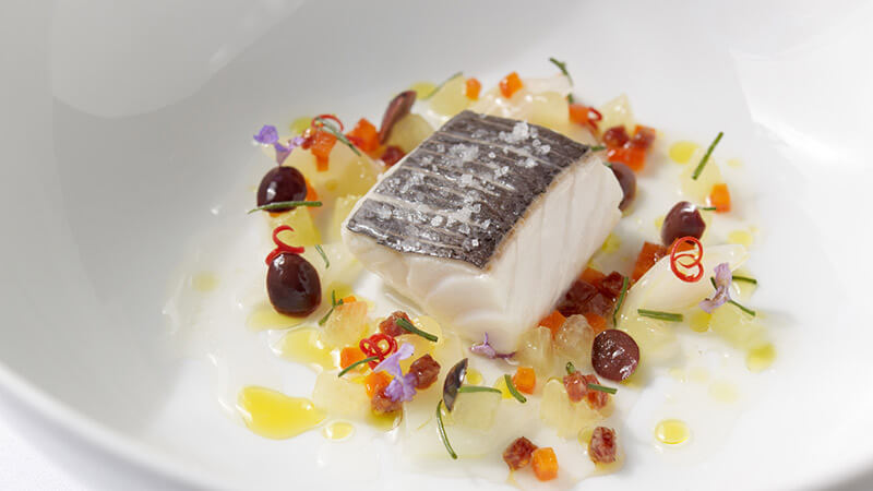 A gourmet plated dish featuring a piece of seabass with crispy skin, accompanied by colorful garnishes and a light sauce, elegantly presented on a white plate.