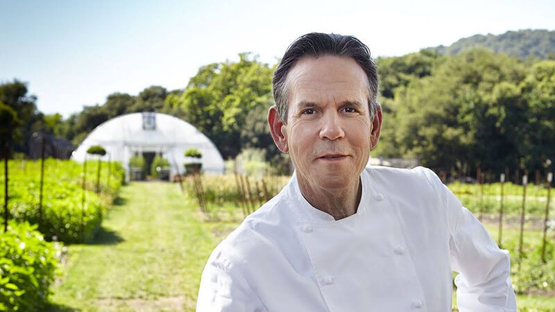 Thomas Keller in a white chef‘s coat standing in front of a lush garden and a greenhouse.
