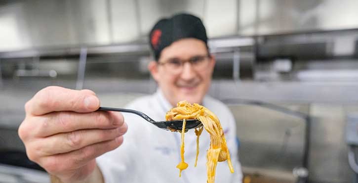 A chef wearing a black cap with a red emblem holds a fork with spaghetti, smiling in a commercial kitchen.