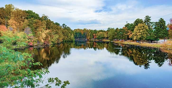 Panoramic view of a serene lake surrounded by lush, colorful autumn trees under a mostly clear sky.
