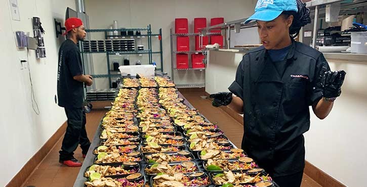 Two chefs carefully arranging prepared meals in containers on a table in a busy kitchen.