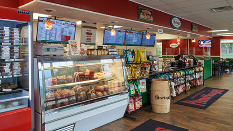 Interior of a Primanti Bros. restaurant showing a deli counter with meats and sandwiches, snack racks, and a menu board above the service area.